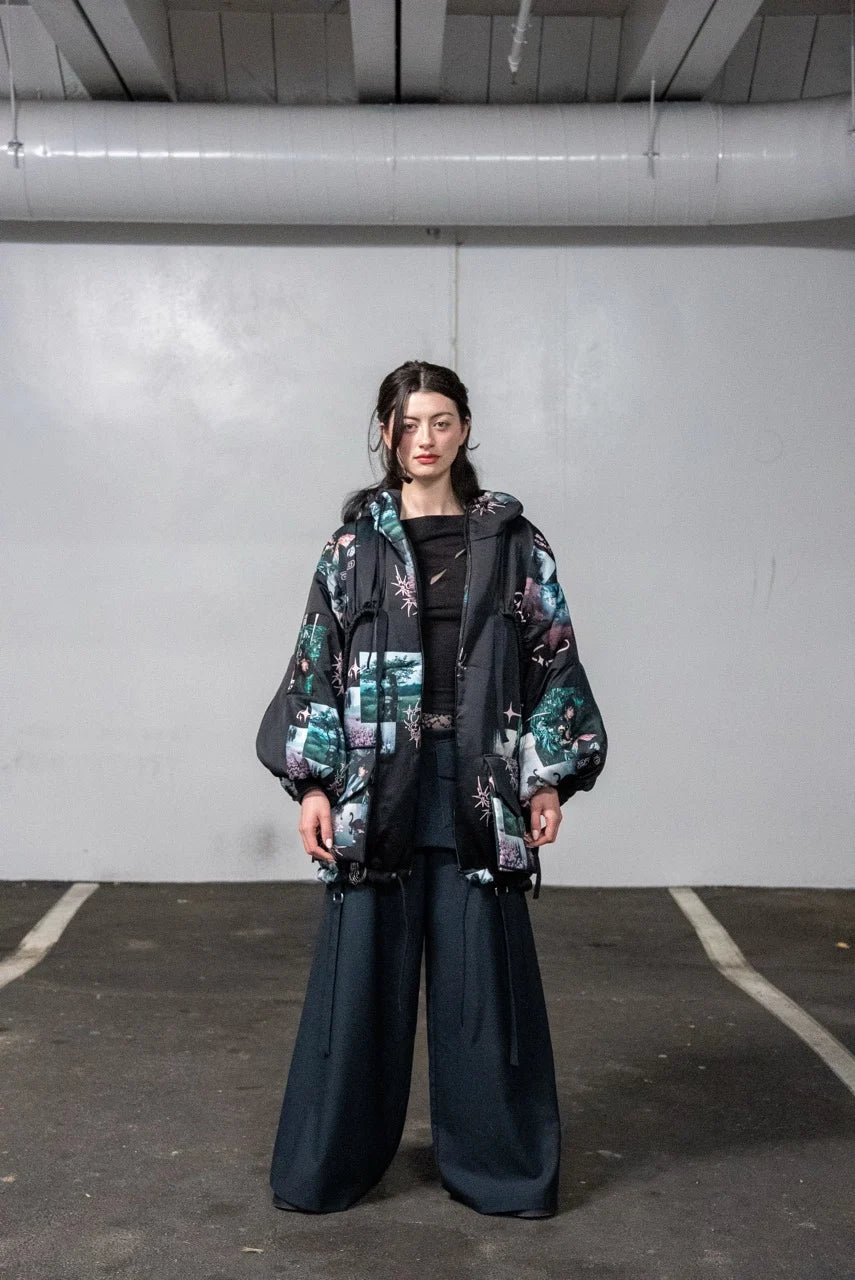 Person wearing a patterned jacket and dark pants standing against a plain wall.